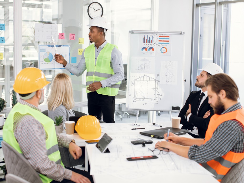 Engineer in a white hard hat and green safety vest presenting charts and construction plans to a team seated around a conference table, with blueprints, laptops, and additional hard hats visible in a modern office setting.